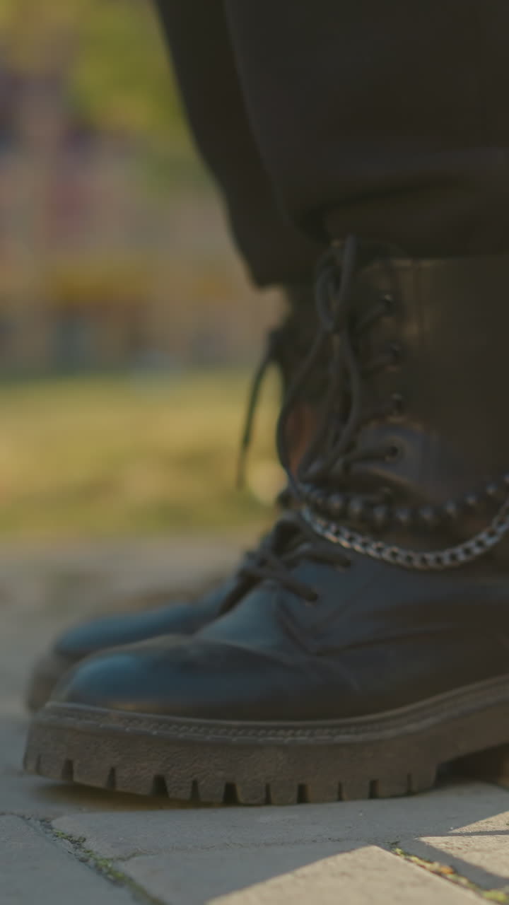 A close-up shot of black boots stepping on a sunlit paved path in a park. The focus is on one boot, with the background softly blurred, highlighting the outdoor setting with hints of greenery