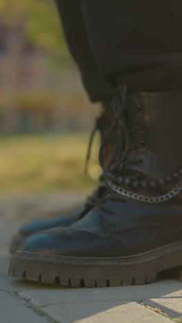 a close-up shot of black boots stepping on a sunlit paved path in a park. the focus is on one boot, with the background softly blurred, highlighting the outdoor setting with hints of greenery