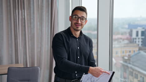 businessman in black shirt, trousers and glasses holding a paper with chart. man following the graph, explaining it to camera.