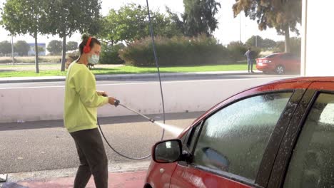 woman cleaning hood of a red car with pressure hose and matching sweatshirt with green hair and background