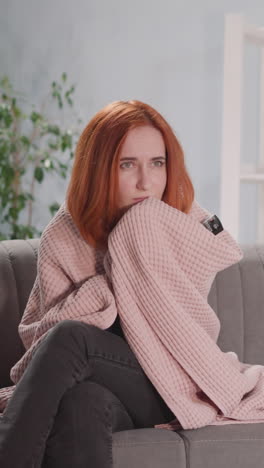 upset woman covered with pink blanket watches dramatic movie on tv. housewife with red hair worries for characters sitting on sofa at home closeup