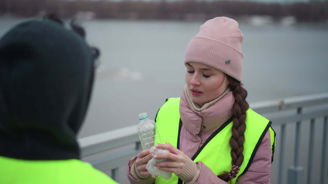 Female worker wearing pink knit hat and reflective vest holding sandwich and bottle of water, standing outdoors on cold cloudy day near waterfront, speaking to colleague during break