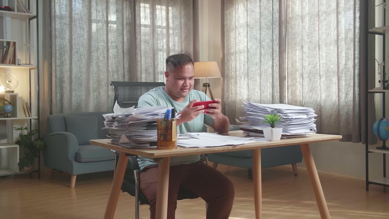 Happy Fat Asian Man Celebrating Winning Game On Smartphone After Working With Documents At The Office