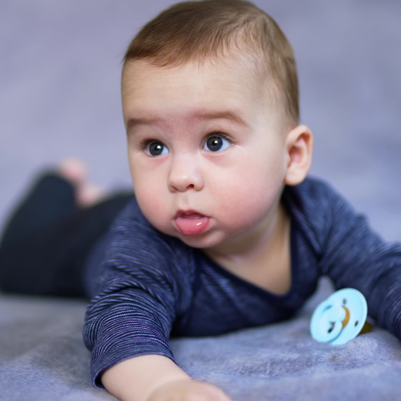 Calm Caucasian baby boy lies on bed with pacifier nearby. Portrait of a beautiful child at grey backdrop