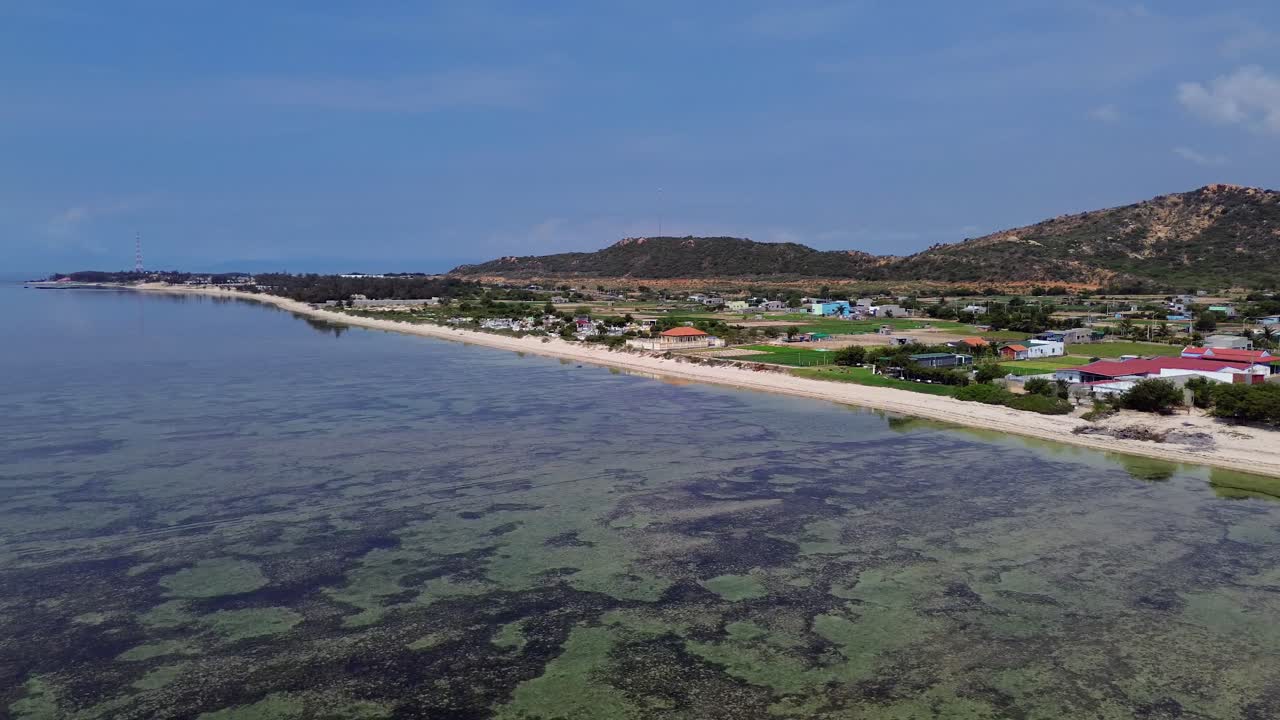 drone flies sidewards from left to right, revealing the beach on the right and houses at the beach, distant mountains, and a clear horizon in Vietnam