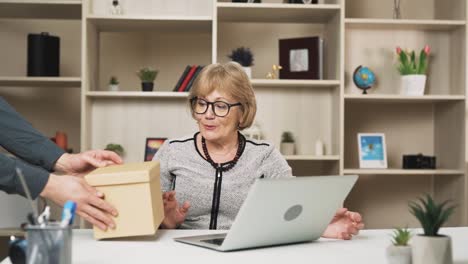 una mujer mayor presiona un botón en su computadora portátil y rápidamente recibe un paquete en una caja, abriéndolo con alegría y agradable sorpresa
