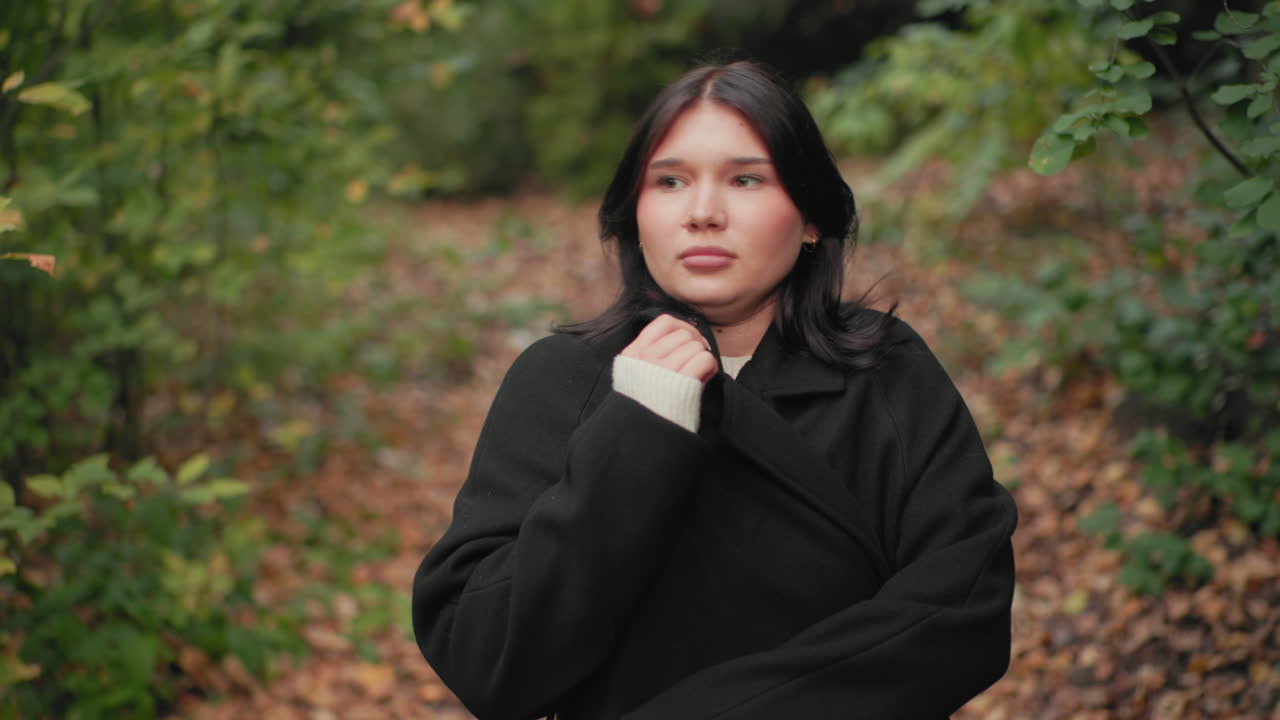 소녀가 숲길을 거닐면서 담요로 몸을 감싸고, 쌀쌀한 날씨에 코트를 단단히 여미고 있어요. 부드러운 가을 낙엽이 발밑에 있고, 시선은 한 곳에 집중되어 있으며, 차분하게 천천히 걷고, 야외에서 계절이 바뀌고 있네요.