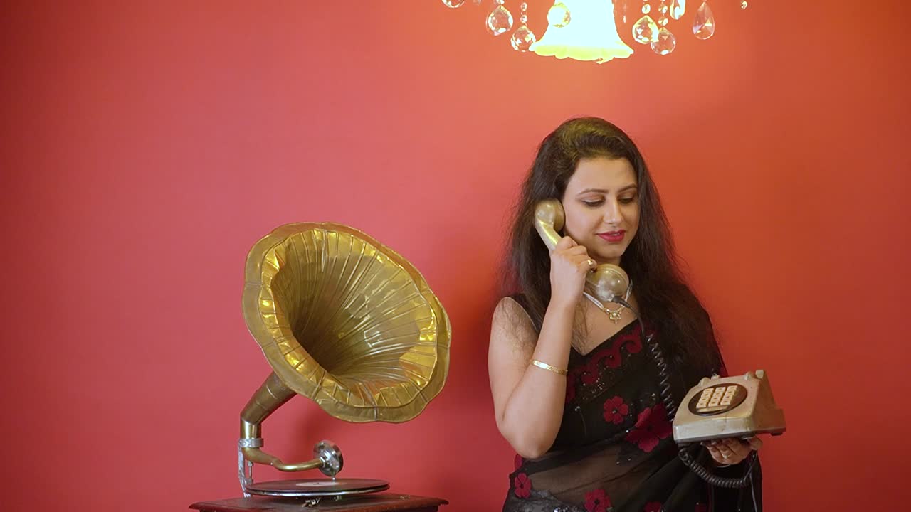 검은색 사리를 입은 인도 여성이 축음기 옆에 서서 금색 빛이 위에 있는 고립된 빨간색 배경에서 전화 통화를 하고 있습니다.