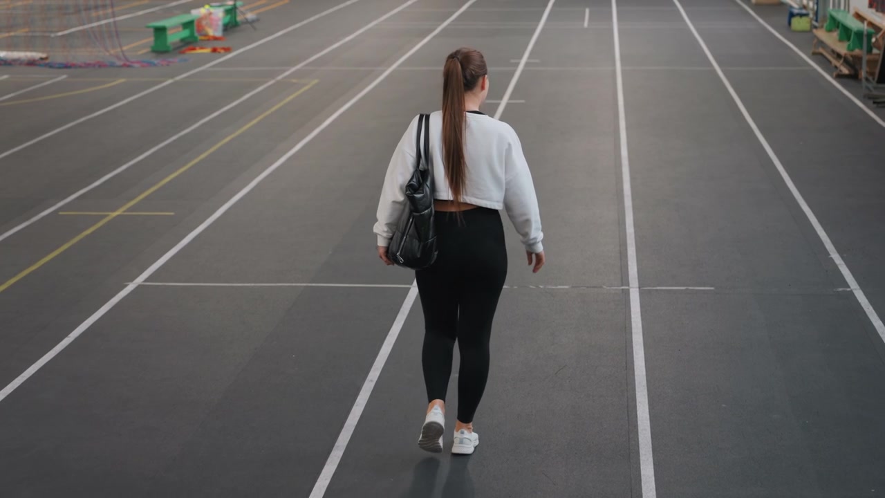Caucasian woman walking away indoor track purposeful pace with long ponytail and gym bag slung over shoulder, heading toward courts and warmup area empty lanes and soft echoes create focused