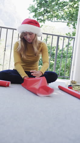 Vertical video of biracial woman wearing santa hat packing christmas presents at home, slow motion
