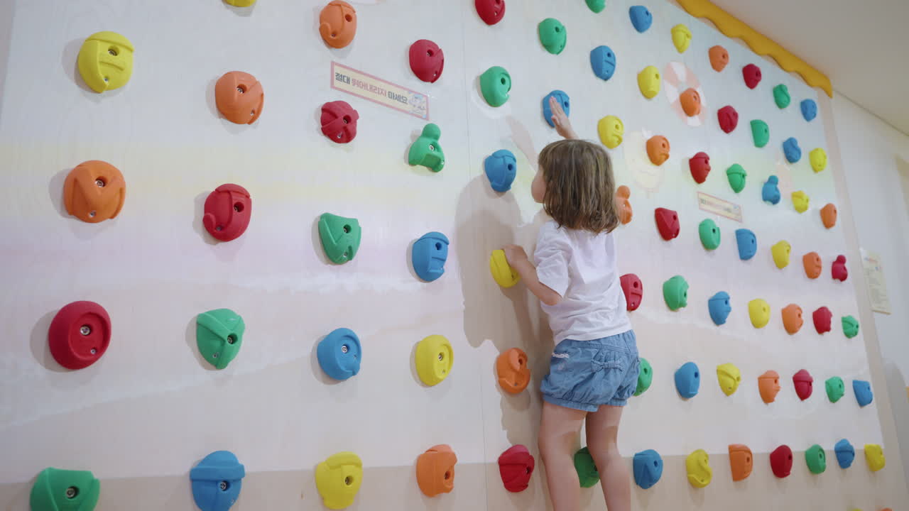 niña activa se sube a la pared de escalada con empuñaduras para golpear objetos de imagen proyectados - jugando a un juego táctil interactivo para el desarrollo de los niños en la sala de juegos interior - tiro de mano