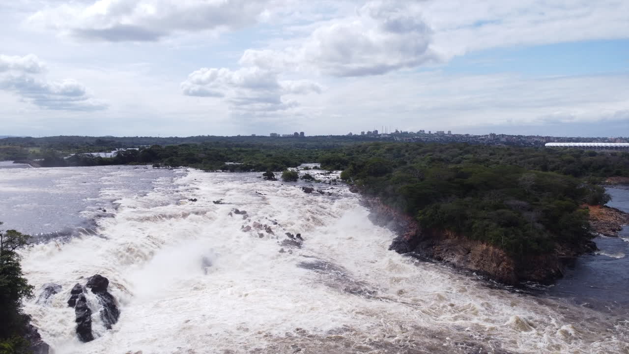 Aerial view of powerful river rapids or waterfall with lush green banks and a distant city skyline under a cloudy sky