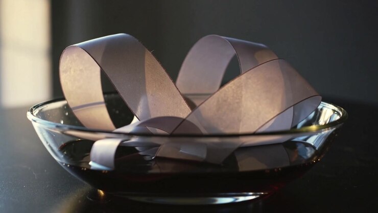 Shifting paper loops in clear glass bowl on dark table tilting and reclining, light causing ripples