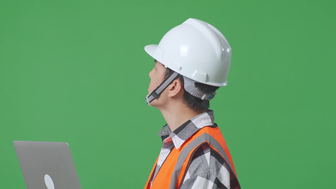 Close Up Side View Of Asian Male Engineer With Safety Helmet Working On A Laptop And Looking Around While Standing In The Green Screen Background Studio