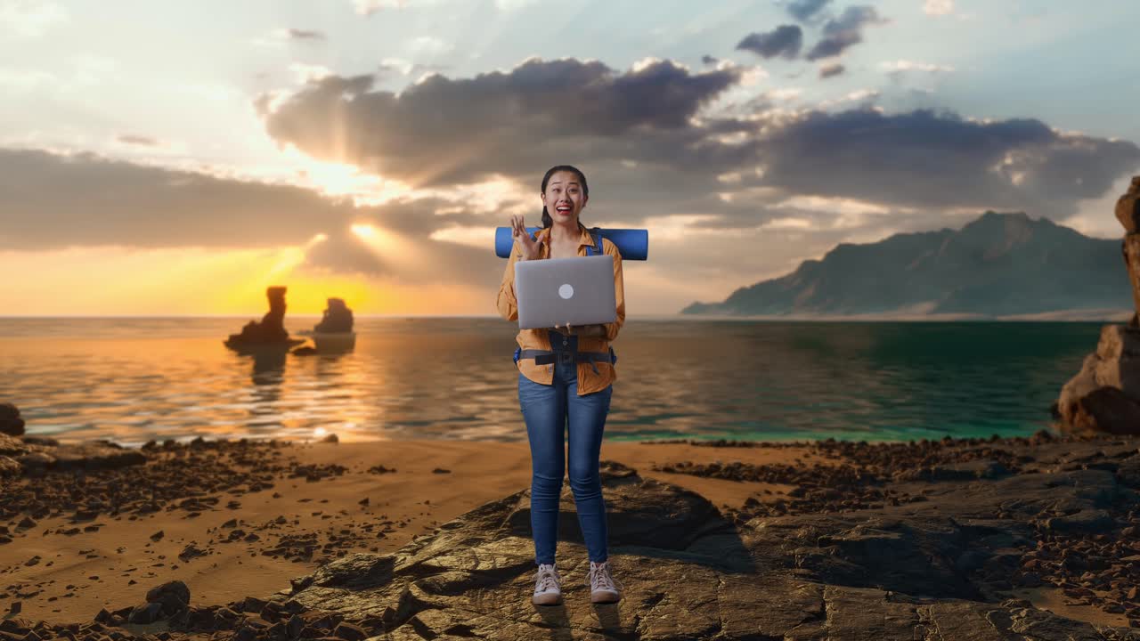 Corpo intero di escursionista asiatica con zaino da alpinismo che guarda un laptop ed esclama "Wow" mentre si trova in spiaggia al tramonto