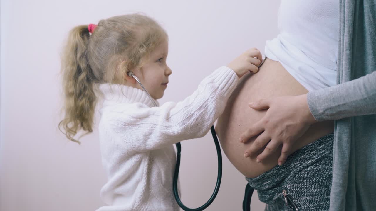 una ragazza divertente mette lo stetoscopio sulla pancia di una mamma incinta nella stanza.