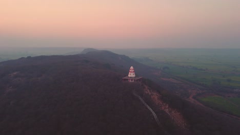 Toma-Aérea-De-Un-Templo-Hindú-En-La-Cima-De-Una-Colina-Con-Escaleras-Que-Conducen-A-él-Durante-La-Puesta-De-Sol-En-Un-Pueblo-De-Gwalior-En-Madhya-Pradesh,-India