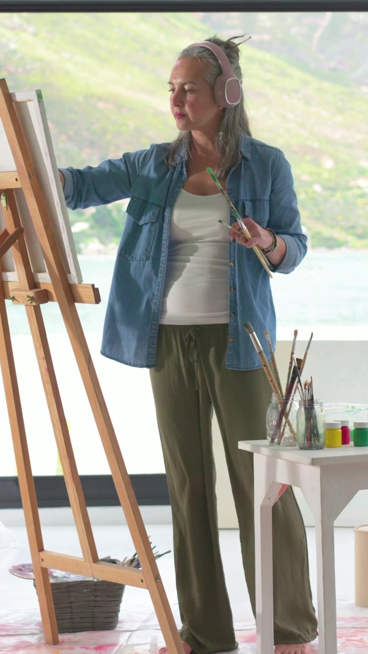 Video verticale: Donna che dipinge con le cuffie, sceglie i pennelli e mescola la pittura sul cavalletto in riva al lago.