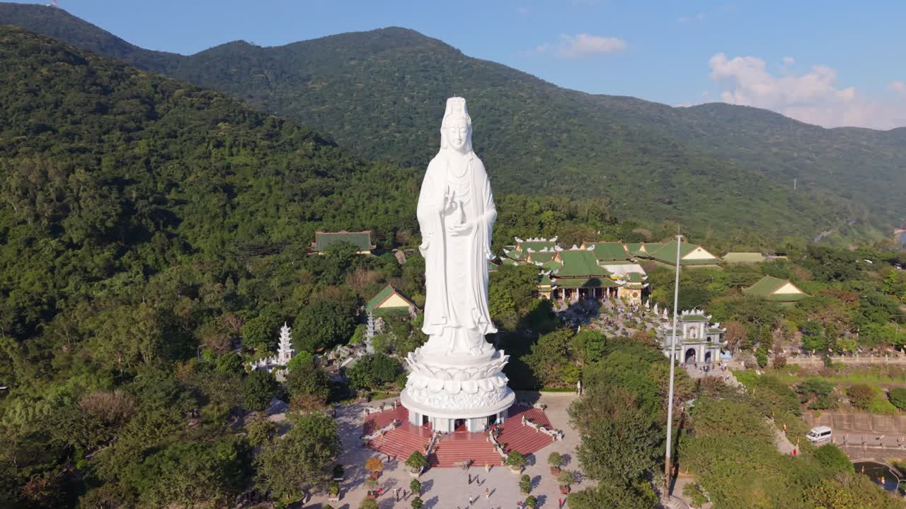 Màn hình dọc: Tượng Phật Bà Trắng Đà Nẵng Việt Nam đứng trên bệ sen trên đỉnh đồi nhìn ra những ngọn núi xanh tươi và khu phức hợp chùa, cảnh quay drone quét góc rộng điện ảnh lúc bình minh