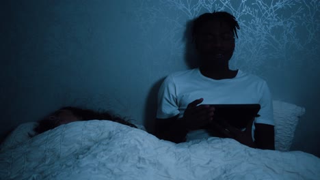 man yawning while watching a tablet device in bed late at night