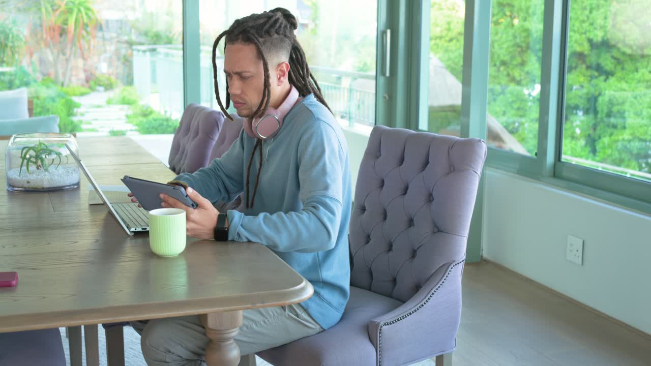 Leaning forward picking up tablet, male tapping screen and typing on laptop browsing at sunroom