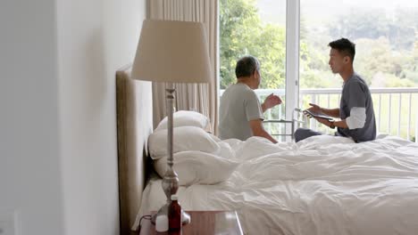 diverse male doctor with tablet discussing with senior male patient in bed at home, slow motion