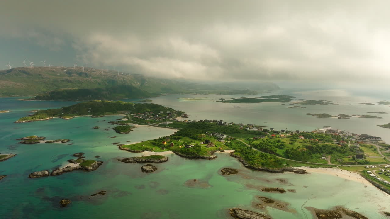 Drone pullback reveals Sommaroy islands and turquoise shallows near Tromsø, with a distant ridge-top wind farm and a broad fjord horizon under soft summer clouds