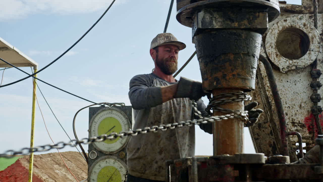 Caucasian man wearing a cap, dirty shirt and protective gloves adjusts the chain around the pipe. Men working at gas or oil production site.