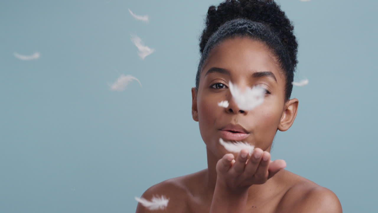 slow motion beauty portrait beautiful african american woman playfully blowing feathers falling touching bare skin enjoying soft skincare gently caressing body in blue background