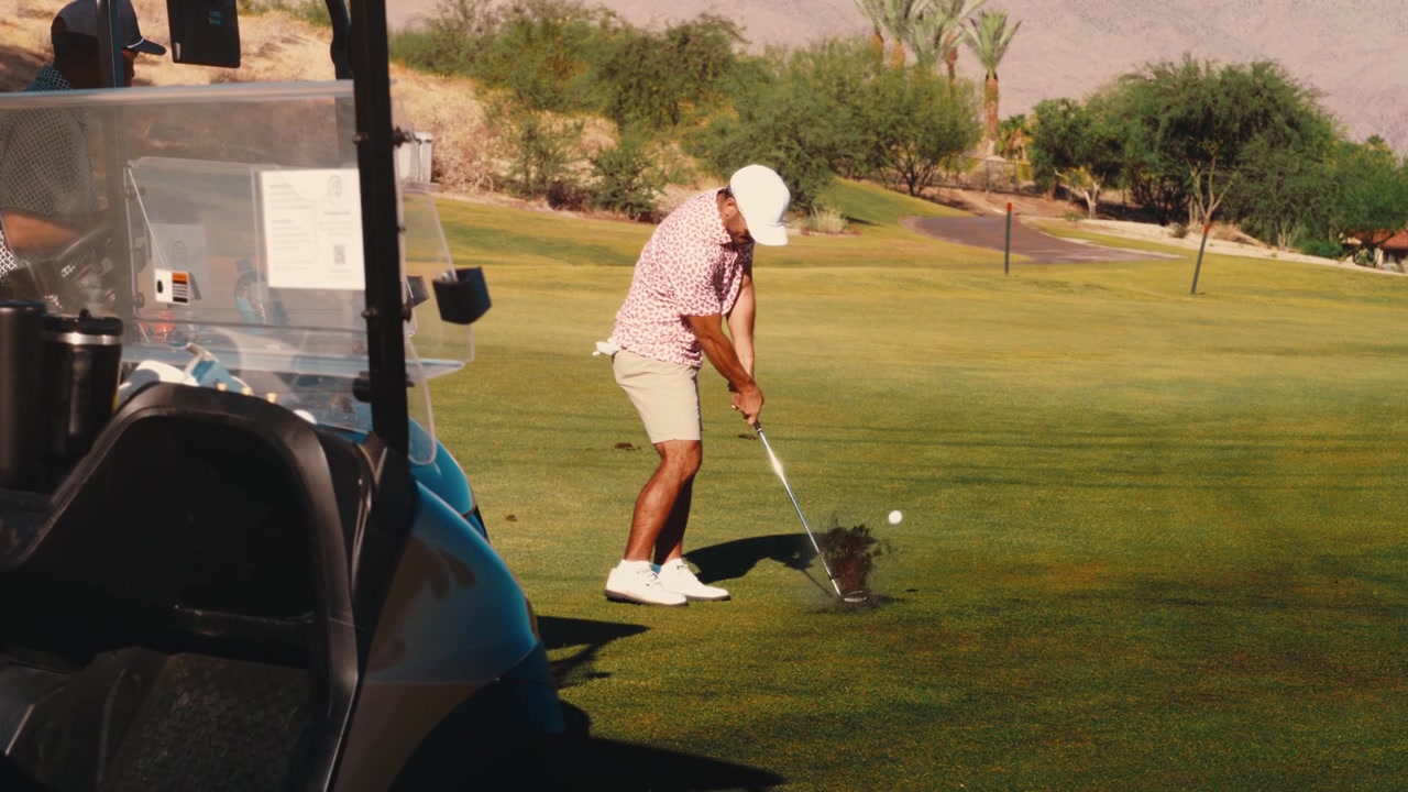 A Black golfer drives through his swing next to a parked golf cart on a sunlit desert fairway, filmed in slow motion. A polished fit for luxury golf and resort ads.