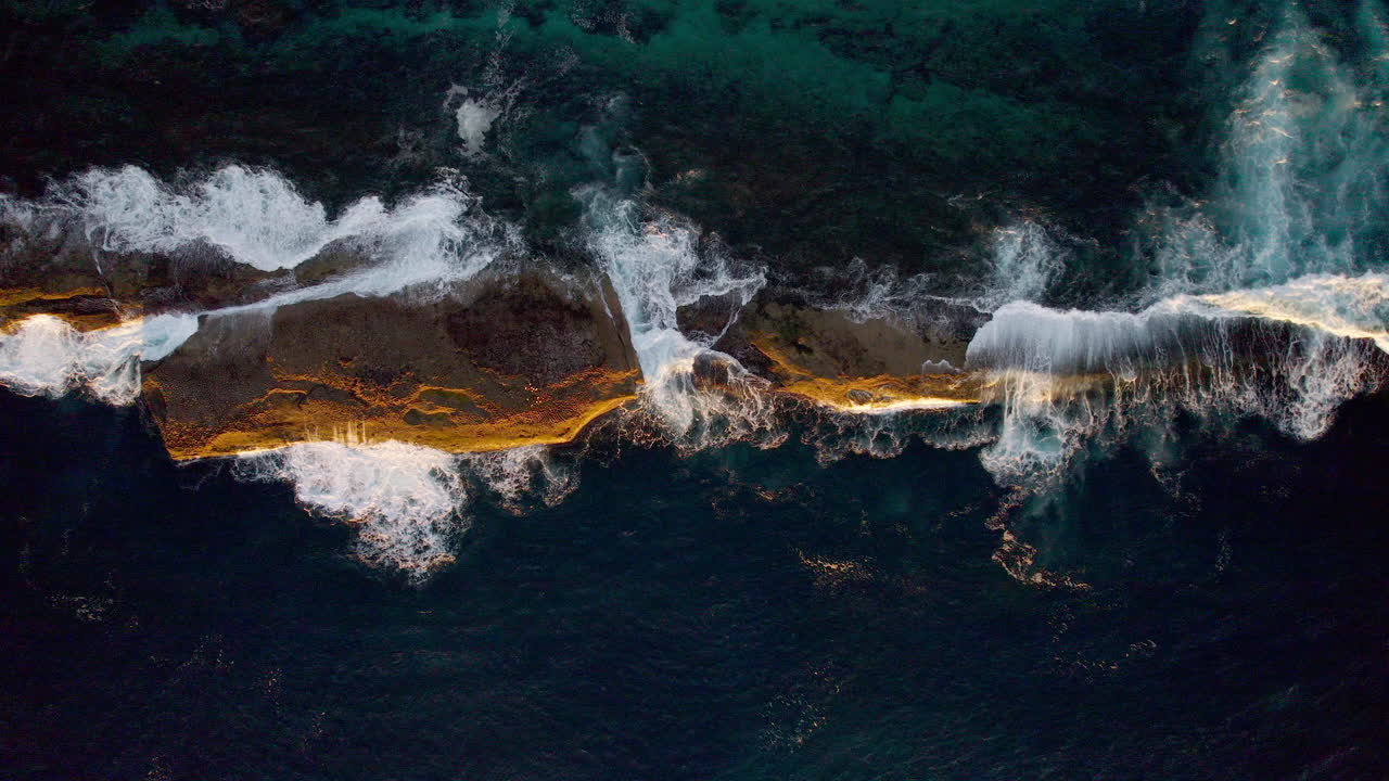 Bathed in golden morning light, a lone rock sits surrounded by shimmering sea. Gentle waves swirl around it, creating a calm and timeless coastal moment near Warden Head, Ulladulla, NSW, Australia.