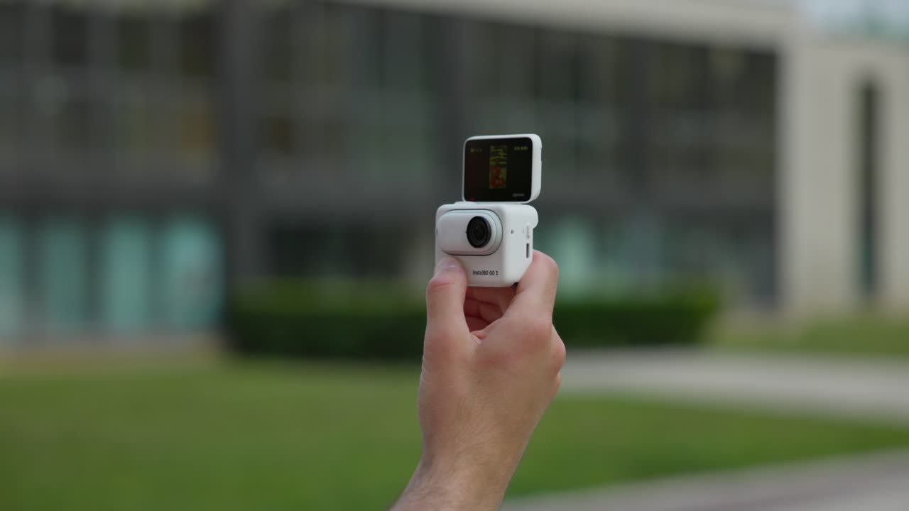la mano izquierda se ve sosteniendo la cápsula de acción insta360 go 3 en modo selfie afuera en un parque urbano