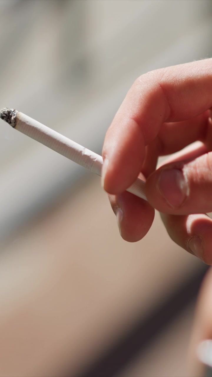 Primer plano de una mano sosteniendo y encendiendo un cigarrillo. La persona manipula un objeto cilíndrico blanco, revelando detalles de textura mientras se enciende con humo visible. Una luz natural suave ilumina la escena.
