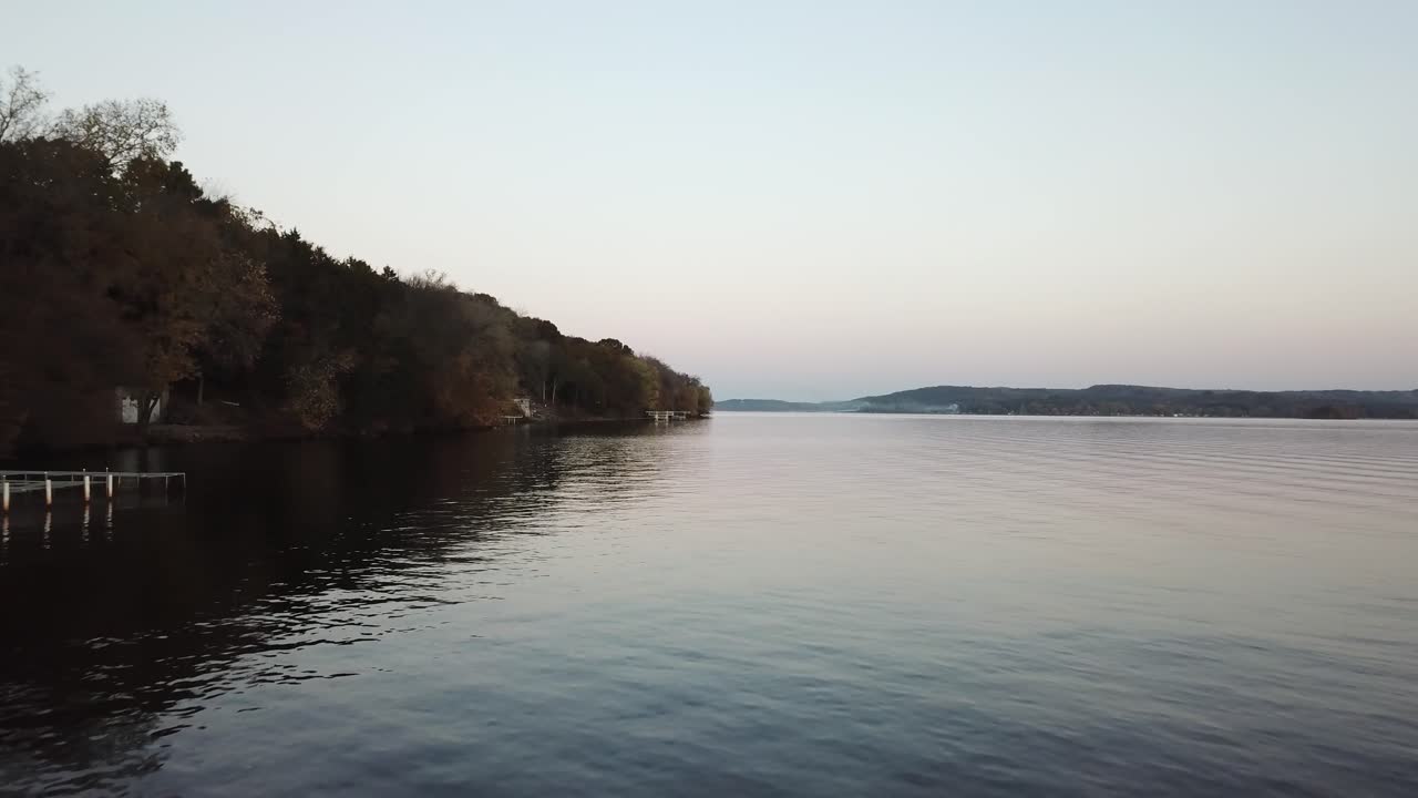 A peaceful lake stretches into the distance under a fading twilight sky. The water is still and reflective, bordered by forested banks with gentle hills in the background.