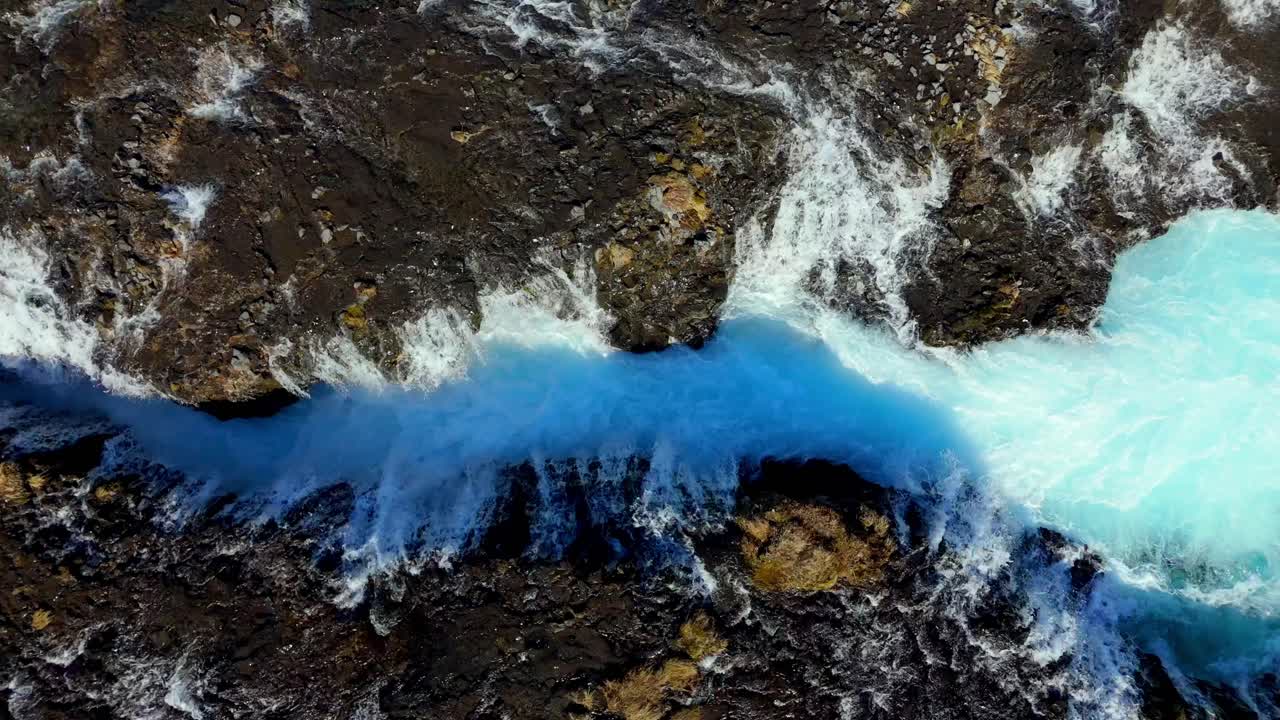 aerial top down of Brúarfoss waterfall in Iceland, featuring vibrant turquoise waters flowing through rugged volcanic terrain in a stunning natural landscape