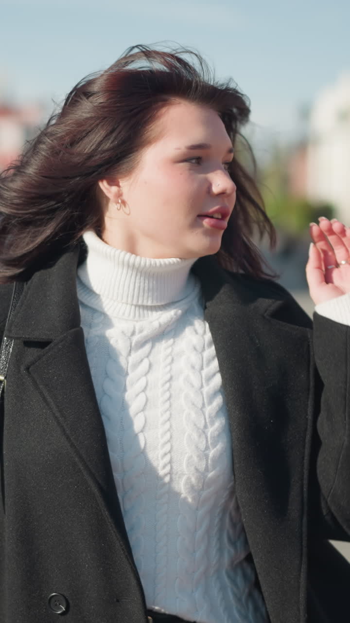 색 케이블 뜨개질 스웨터와 검은 코트를 입고 왼쪽으로 바라보는 머리를 조정하는 젊은 여성, 빛 도시 거리에서, 캐주얼 우아함을 발산