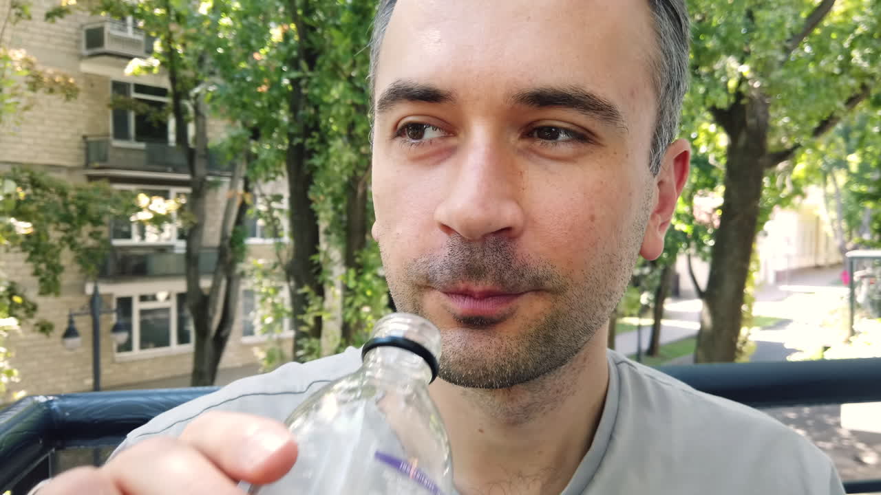 A man takes a sip of a chilled beverage while sitting outside in a lively city park filled with trees. Sunlight filters through the leaves, creating a relaxing atmosphere