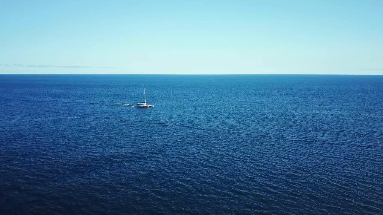 A sailboat gracefully moves across the calm, vast ocean under a clear blue sky, creating a serene and tranquil maritime scene. The expansive sea and horizon evoke a sense of freedom