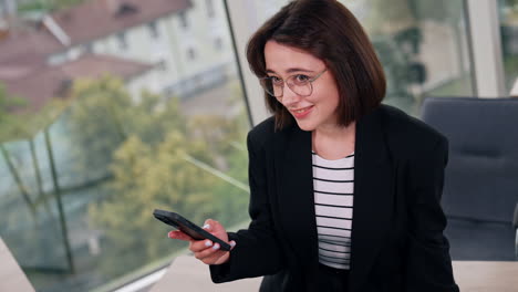 positive smiling lady wearing glasses and black jacket uses her smartphone. high angle view on the young woman surfing internet on the phone.