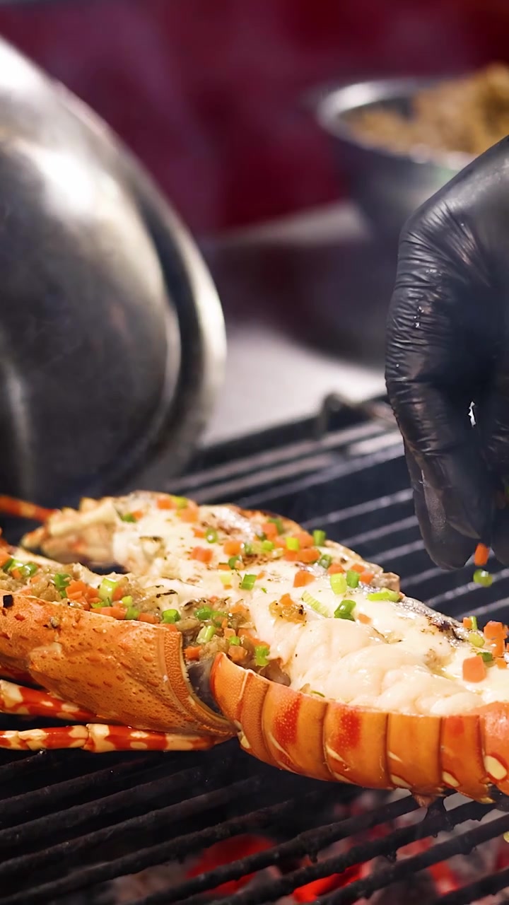 Gloved chef sprinkles diced vegetables onto lobster tail grilling over open flame, vibrant street setting