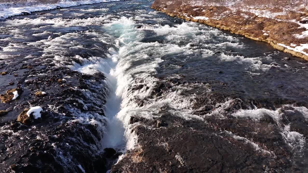 Hyperlapse view of Brúarfoss waterfall, Iceland, showcasing its rapid flow and vibrant turquoise water against a snowy backdrop.