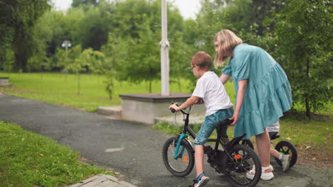 a mother supports her older son as he learns to ride a bicycle while the younger son propels himself forward using his legs on a balance bike