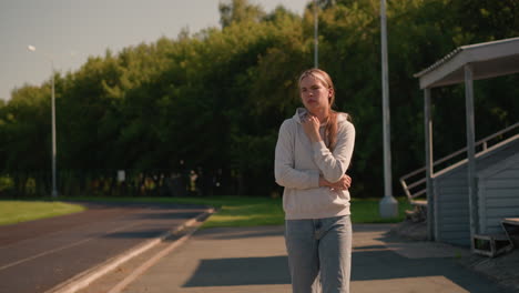 young woman in casual hoodie walks thoughtfully near an empty stadium on a bright sunny day, surrounded by lush green trees, background features empty bleachers and tall electric poles