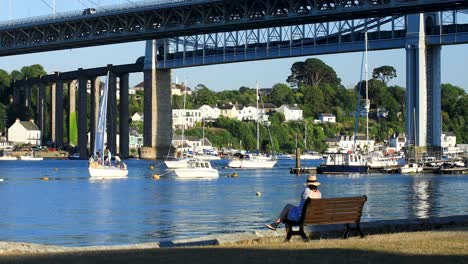 a lady with a hat sits on the bench overlooking the river tamar with the tamar and royal albert bridge in the background on a summers evening in saltash, cornwall