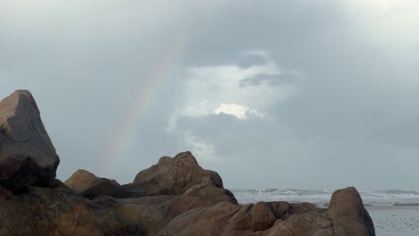 Above-the-tumultuous-sea-waves-crashing-against-Zahara's-rugged-and-rocky-shoreline,-a-rainbow-adorns-the-sky,-epitomizing-the-allure-and-natural-marvels-of-the-coastal-scenery