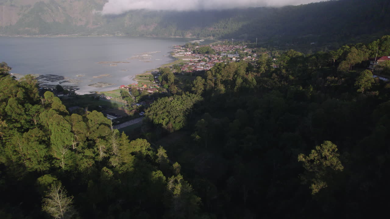 Aerial shot in Bali flying over blue lake ata Mt Batur surrounded by fishertown and a forests on a mountain