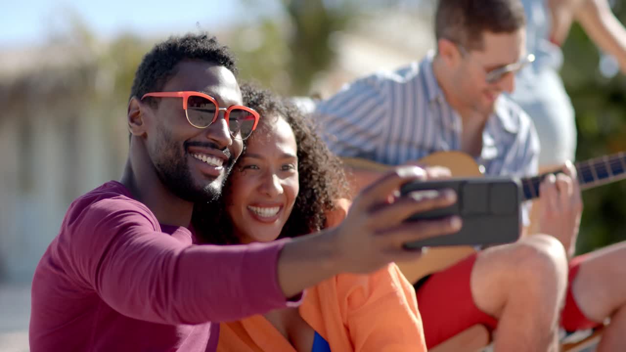 un grupo feliz y diverso de amigos tomando selfies y tocando la guitarra en la playa con la casa de la playa