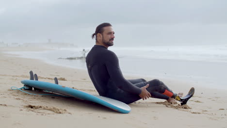 vista lateral de un deportista con una pierna artificial sentado en una playa de arena y descansando después de un entrenamiento de surf en el océano