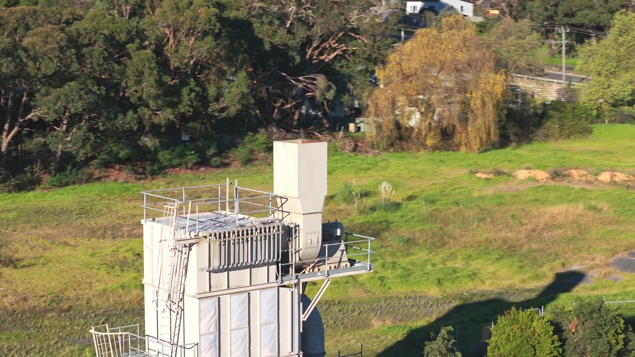 Video drone della Cava di Montrose a Melbourne mostra una prominente torre industriale tra erba rigogliosa, alberi e un cielo azzurro limpido, con la luce del sole che illumina l'ambiente circostante e l'ampio paesaggio