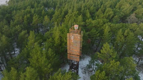 aerial birdseye view of bernati lighthouse surrounded by lush green pine tree forest with light snow, nordic woodland, baltic sea coast, sunny winter day, latvia, wide orbiting drone shot